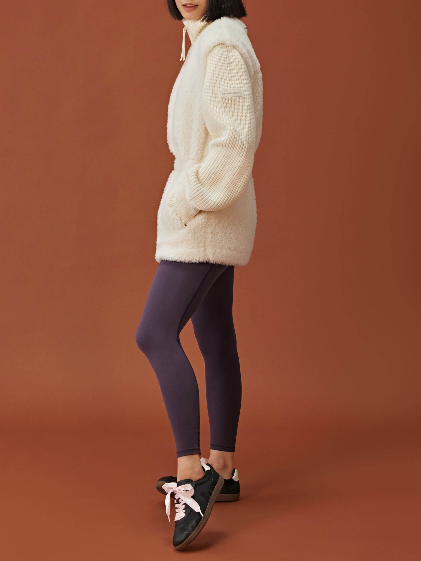 side of woman in white fleece coat with gathered waist and knitted sleeves and dark purple leggings