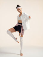 Model wearing white jacket, black shorts, and white leg warmers stretching.
