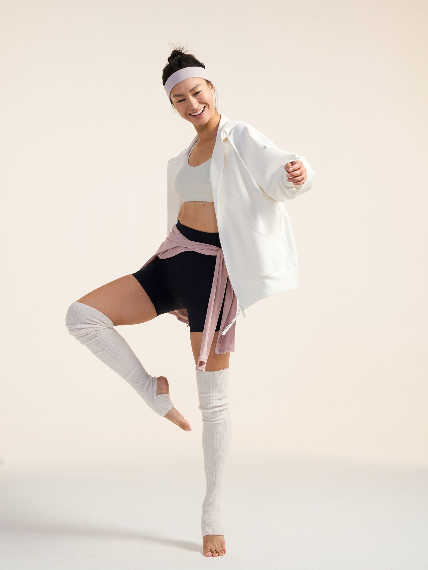 Model wearing white jacket, black shorts, and white leg warmers stretching.