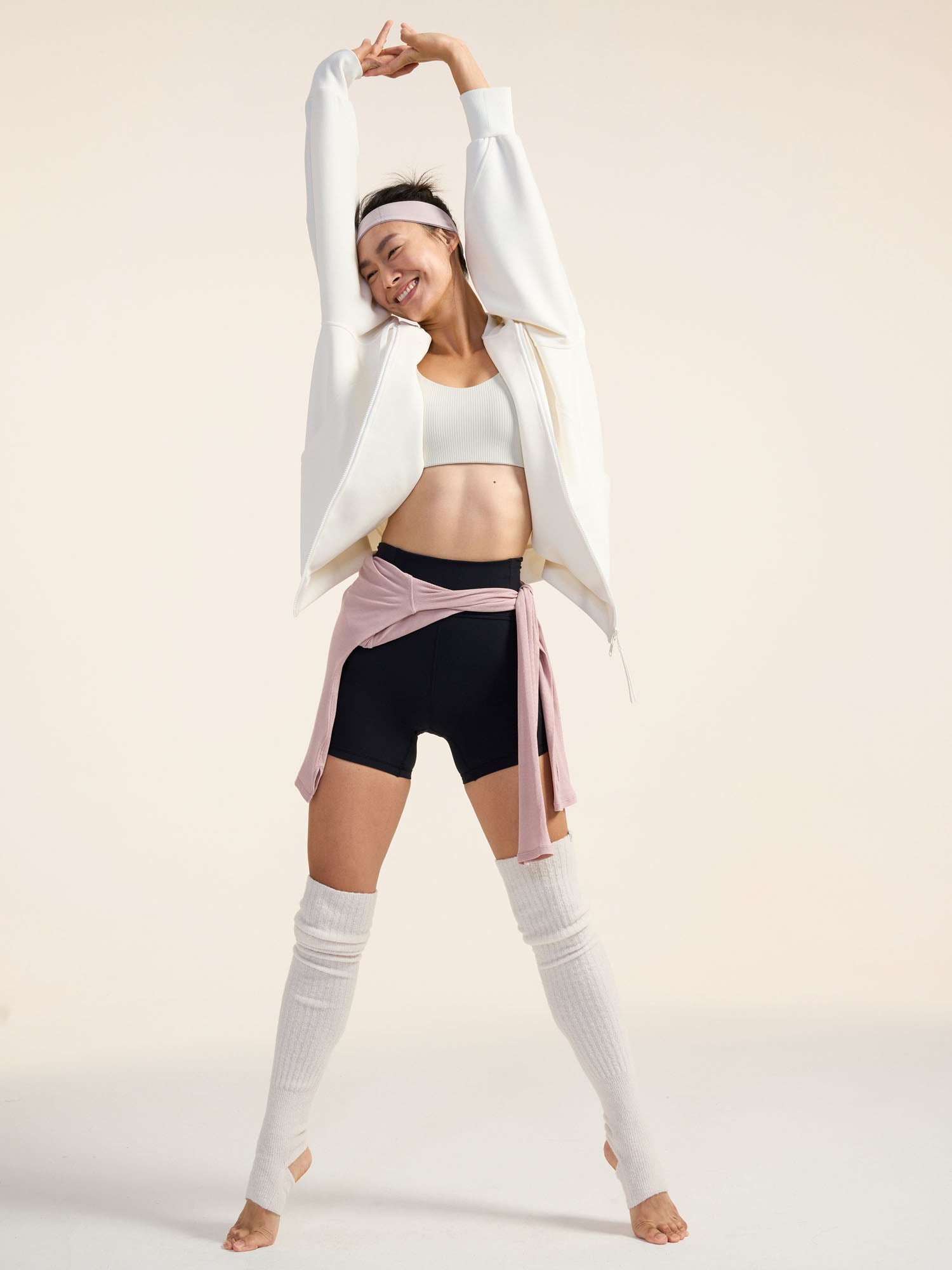 Model wearing white jacket, black shorts, and white leg warmers stretching.