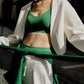 A woman wears a green sports bra, a white sports cardigan, and white sweatpants. There is a building in the background.