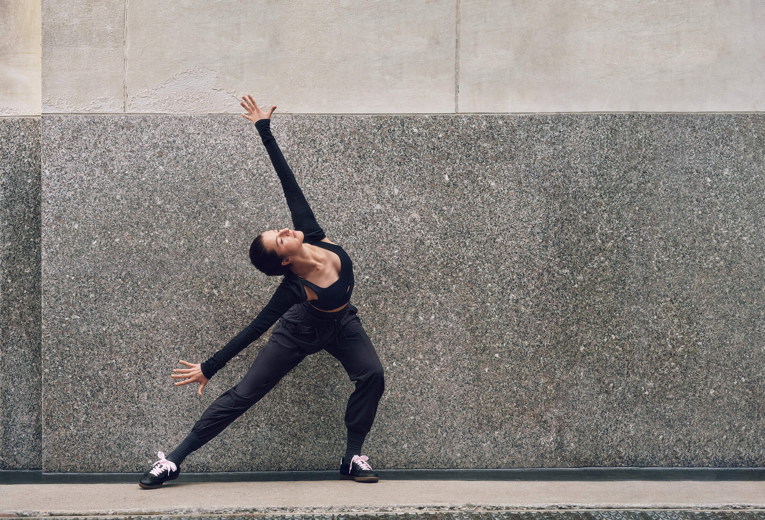 Dancer leaning forward wearing black sports bra, pants, and shrug with arms outstretched