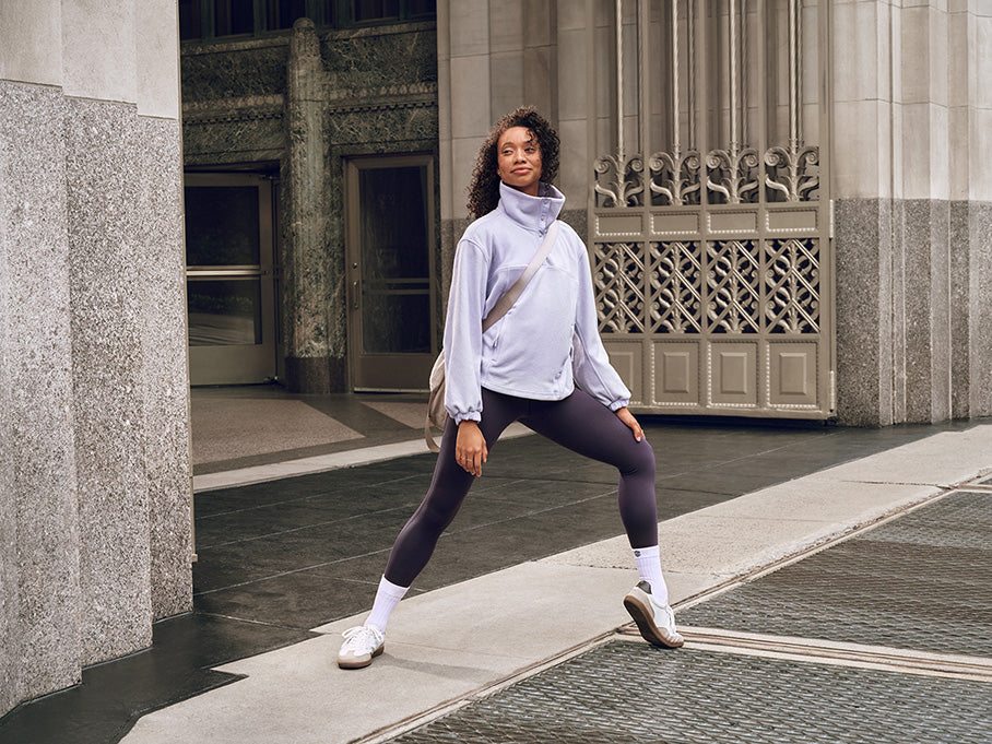 Woman wears light purple sweatshirt, dark purple leggings and gray crossbody bag, standing and pointing one foot.