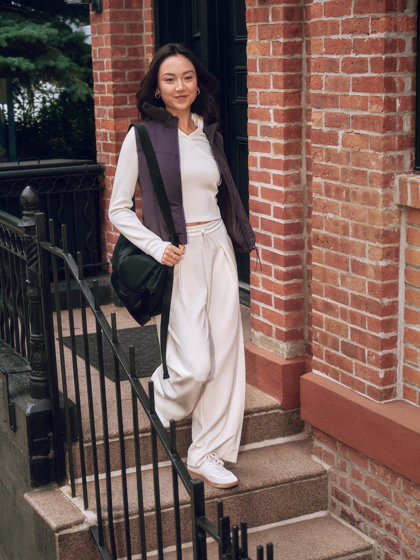 Woman wearing white sweatshirt and sweatpants with purple vest on top, carrying a black bag and walking down the stairs