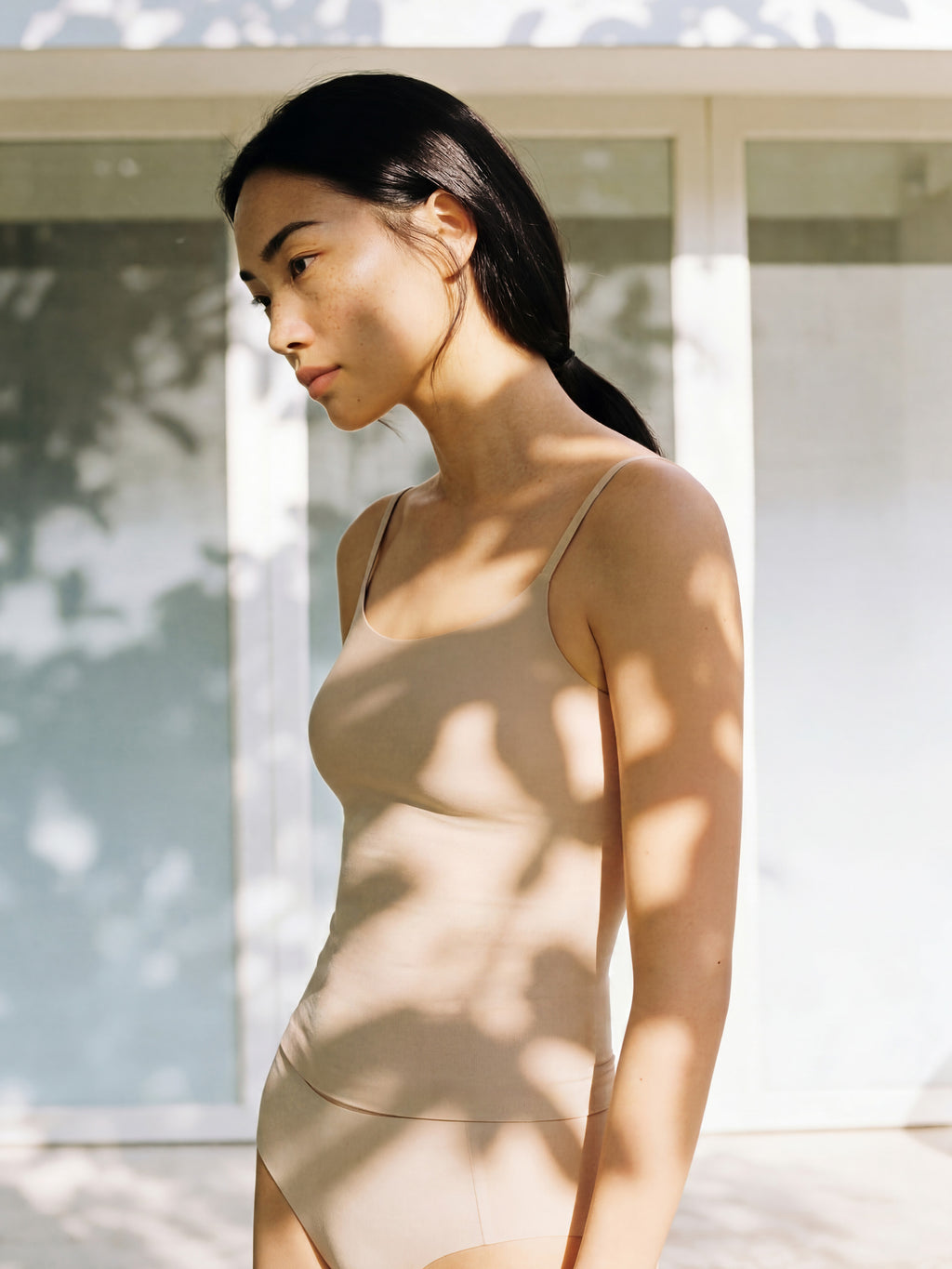 Woman wearing a beige swimsuit standing outdoors with sunlight casting shadows.