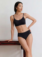 A woman in a matching Barely Zero Anchored Cup Strap Bra and underwear stands with one hand on a red wooden table, gazing to the side against a plain light-colored background. Black color.