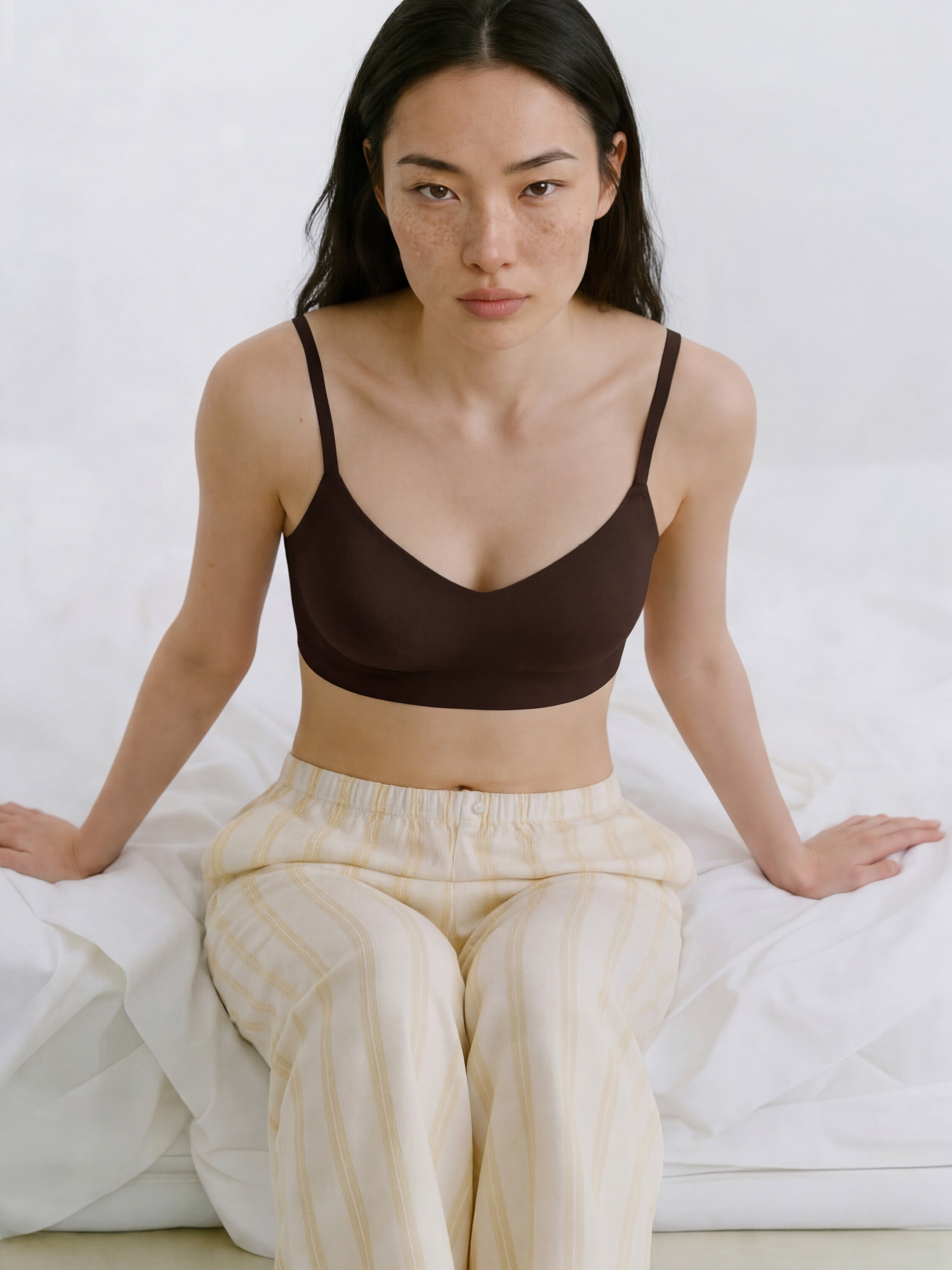 Woman wearing a brown bralette and beige striped pants sitting on a white bed.