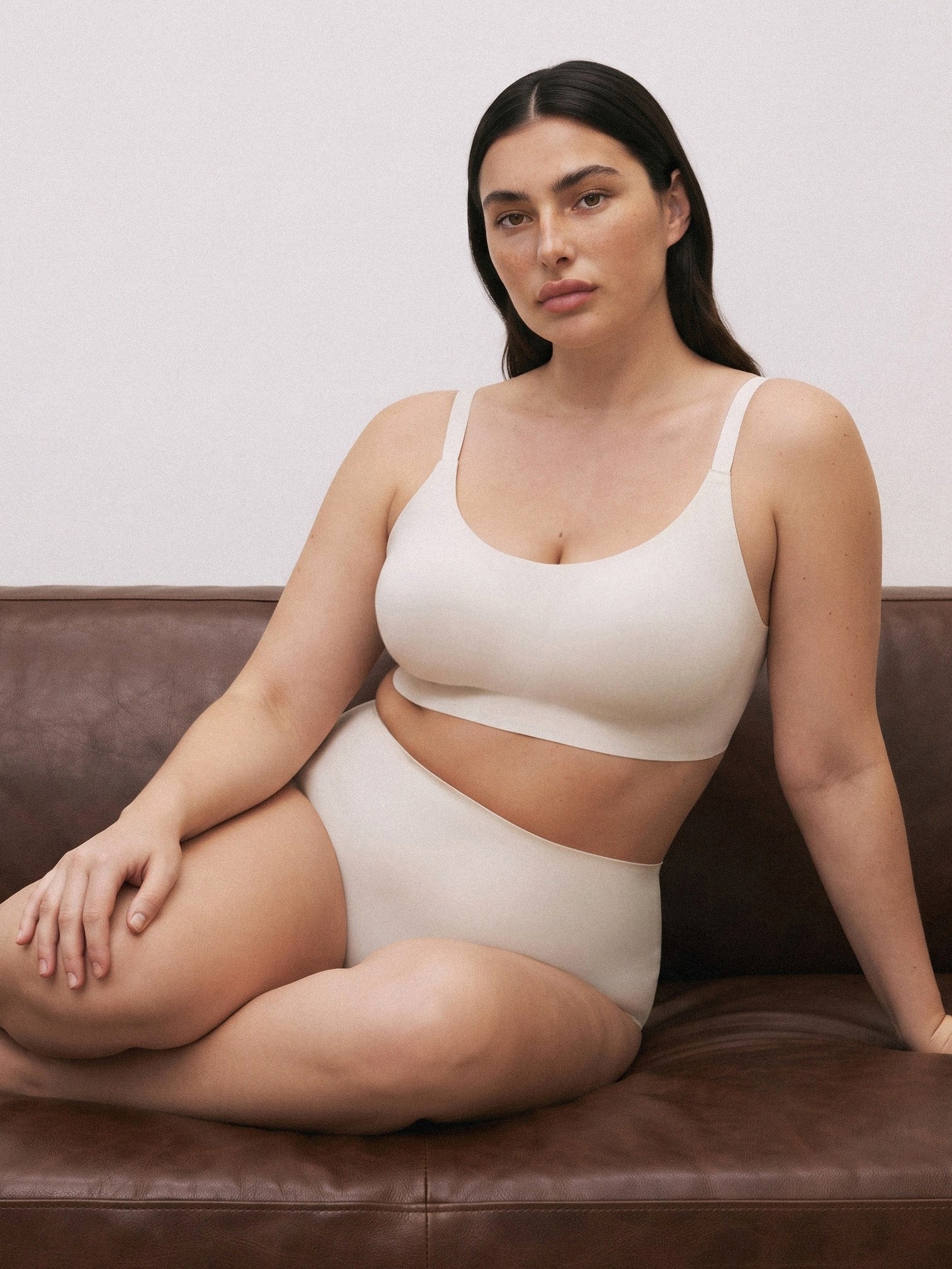 A woman sits on a brown leather couch wearing a matching beige bralette and high-waisted brief set against a plain light wall. Vanilla Bean
