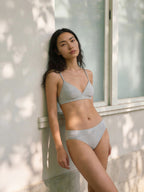 Woman wearing a light gray bikini standing against a white wall with a window.