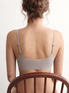 A woman viewed from behind wearing a light taupe-gray bra, seated on a wooden chair against a plain white background. Chateau Grey