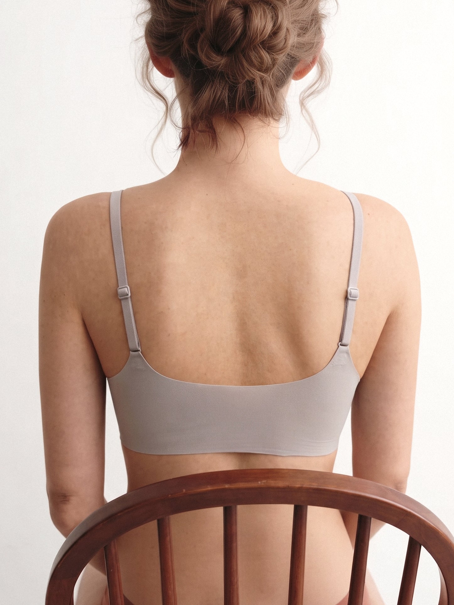 A woman viewed from behind wearing a light taupe-gray bra, seated on a wooden chair against a plain white background. Chateau Grey