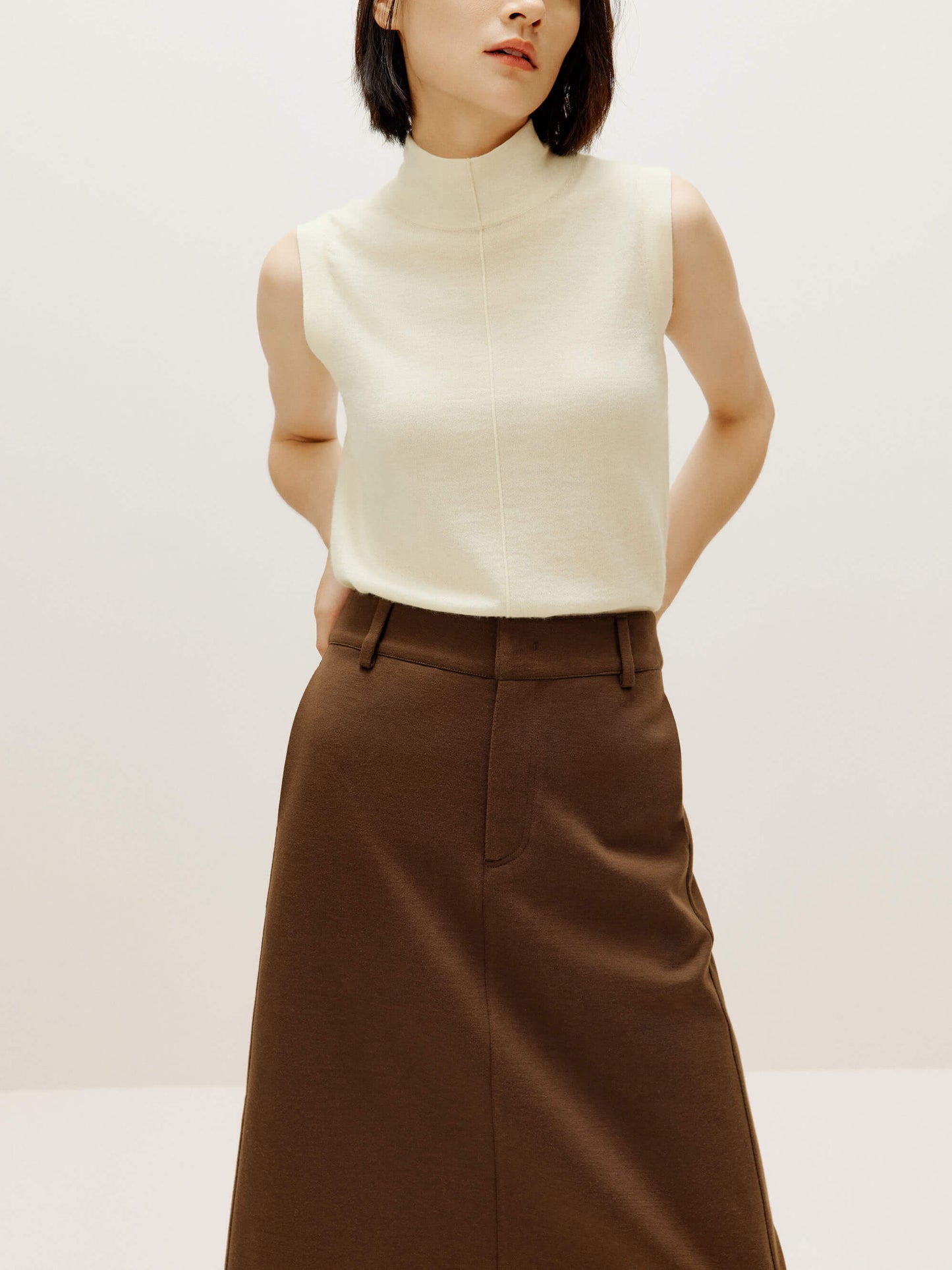 A woman wears a white silky wool mock neck sleeveless sweater and a brown skirt.