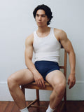 Male model in white tank top and brief seated on wooden chair. Navy color. 