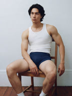 Male model in white tank top and brief seated on wooden chair. Navy color. 