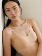 A woman with long dark hair and natural makeup wears the Barely Zero Scallop Clasp Bra, leaning against a light wall. The bra provides breathable comfort and lightweight shaping, shown in soft, natural lighting. Treacly Almond color.
