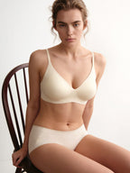 A woman seated on a dark wooden chair wearing a light cream-colored bra and matching high-waisted briefs against a plain light background. Eggwhite