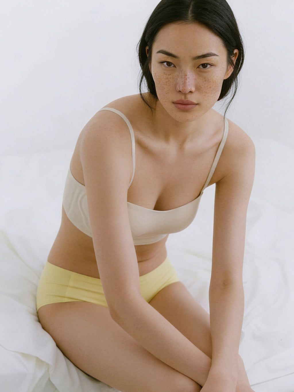 Woman wearing a beige bra and yellow underwear sitting on a white bed. Eggwhite color.