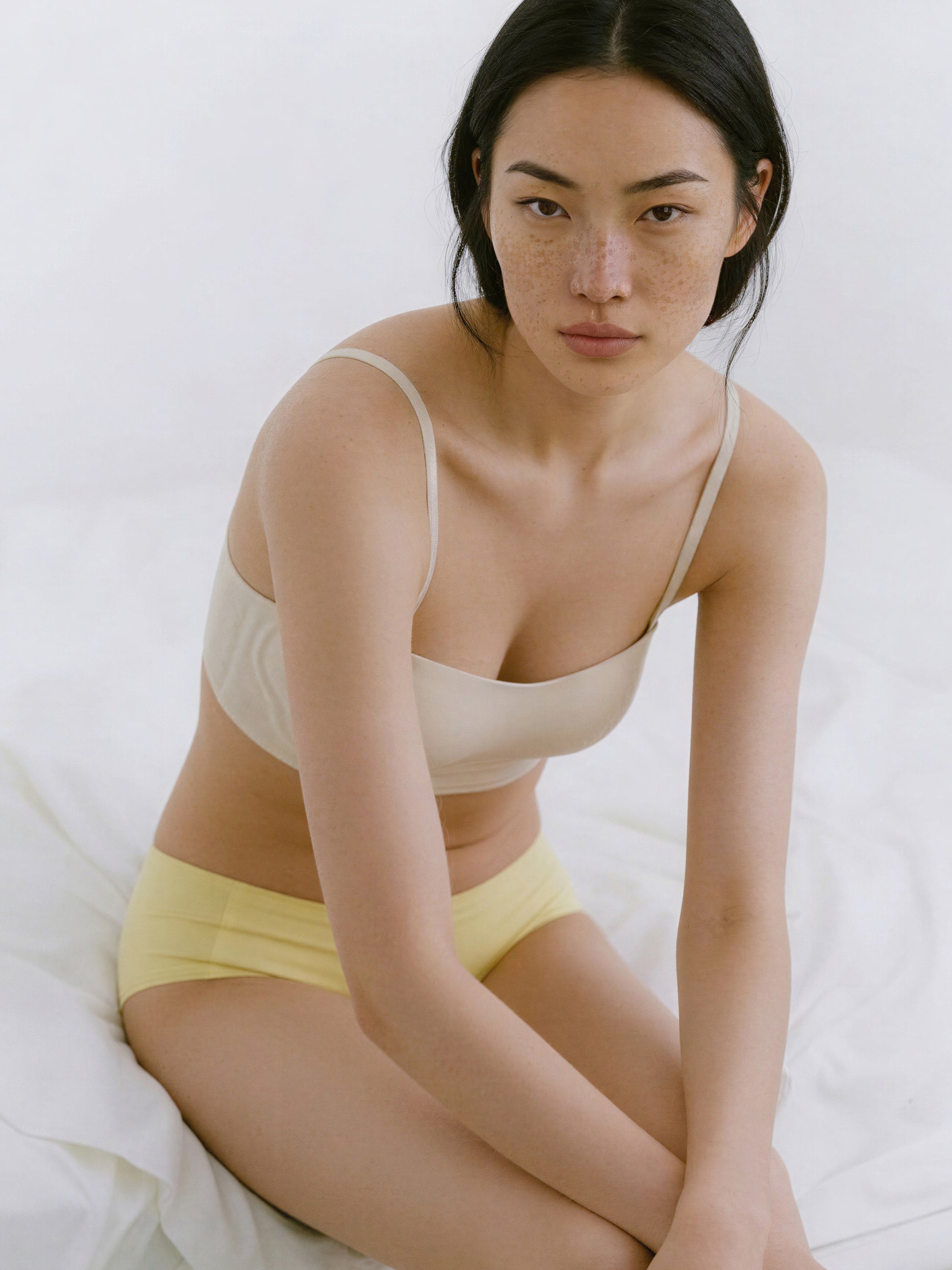 Woman wearing a beige bra and yellow underwear sitting on a white bed.