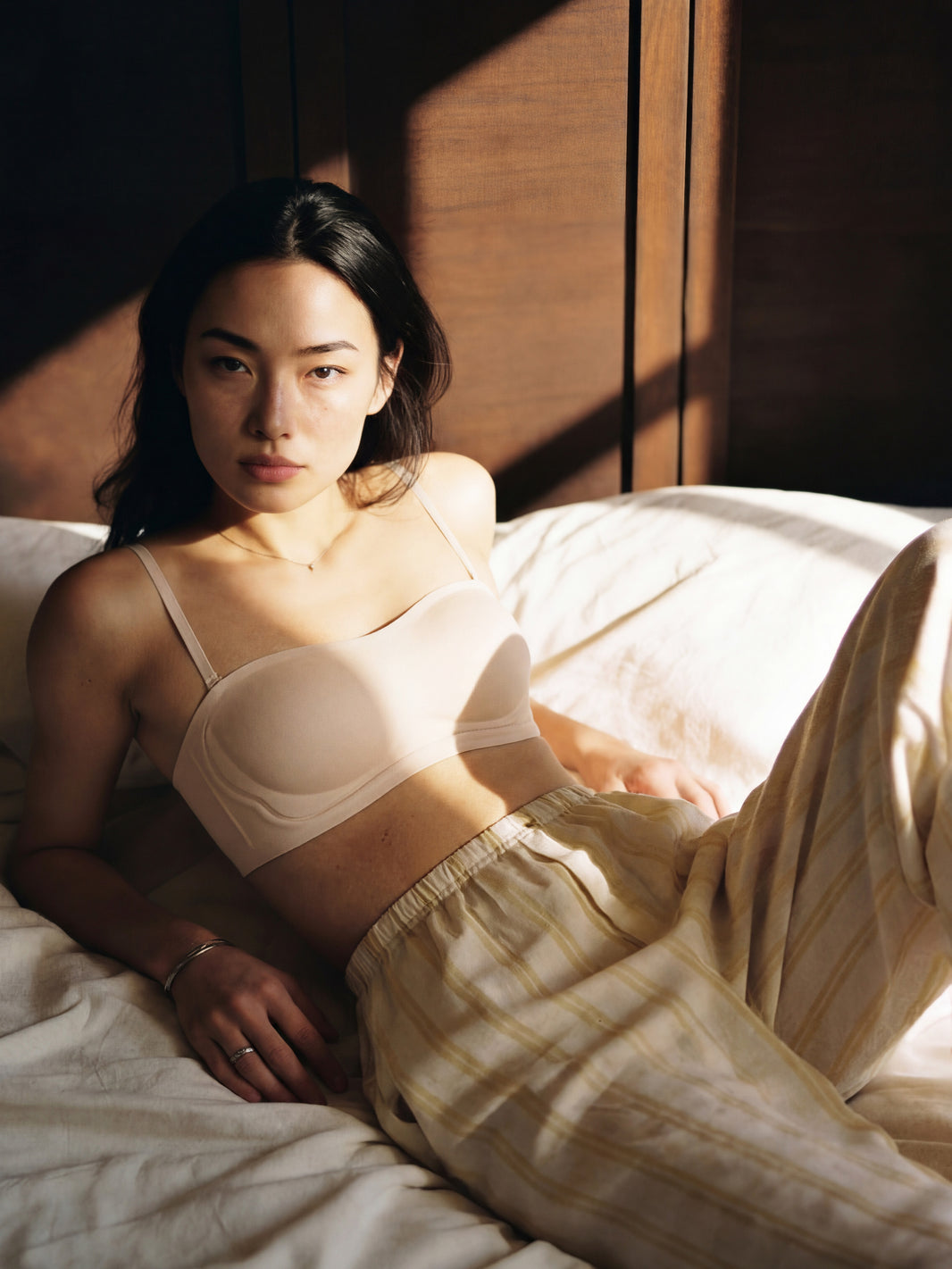 Woman in a white bra and beige pants lying on a bed with soft lighting.