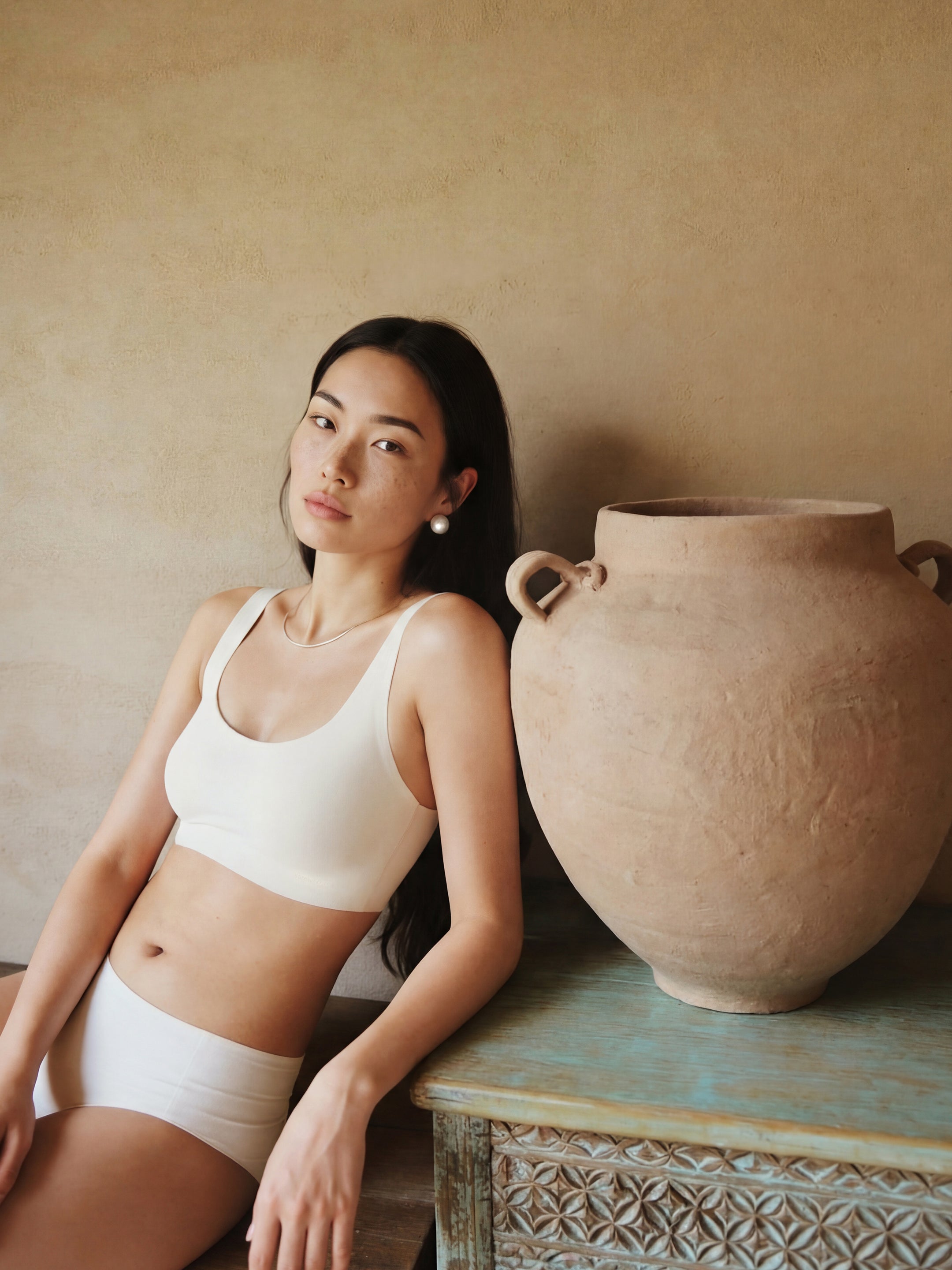 Woman in white bra sitting next to a large terracotta pot against a beige wall.