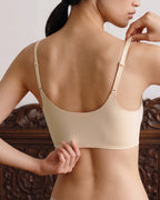 A woman shown from the back wearing a peach pure wireless bra with adjustable shoulder straps, set against a dark carved wooden headboard and a light neutral wall. peach pure