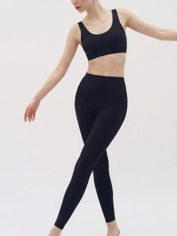 A woman modeling a matching black activewear set consisting of a scoop-neck sports bra and high-waisted leggings, photographed against a clean white studio background.