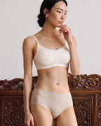 A woman modeling a matching white bralette and high-waisted brief set, standing in front of a carved wooden headboard against a light neutral wall. eggwhite