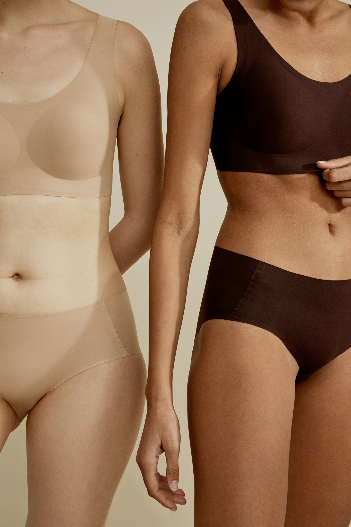 Two women showcasing Barely Zero Classic Bra Trio in neutral colors, one in a beige set and the other in brown, focusing on torso and legs against a plain background.