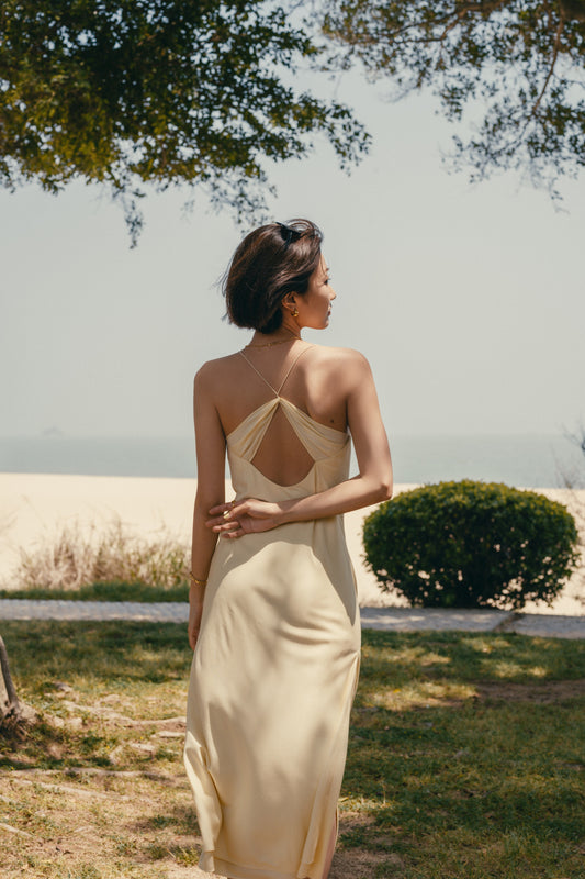 Back view of woman standing on grass wearing cream colored dress with thin straps and cross back design