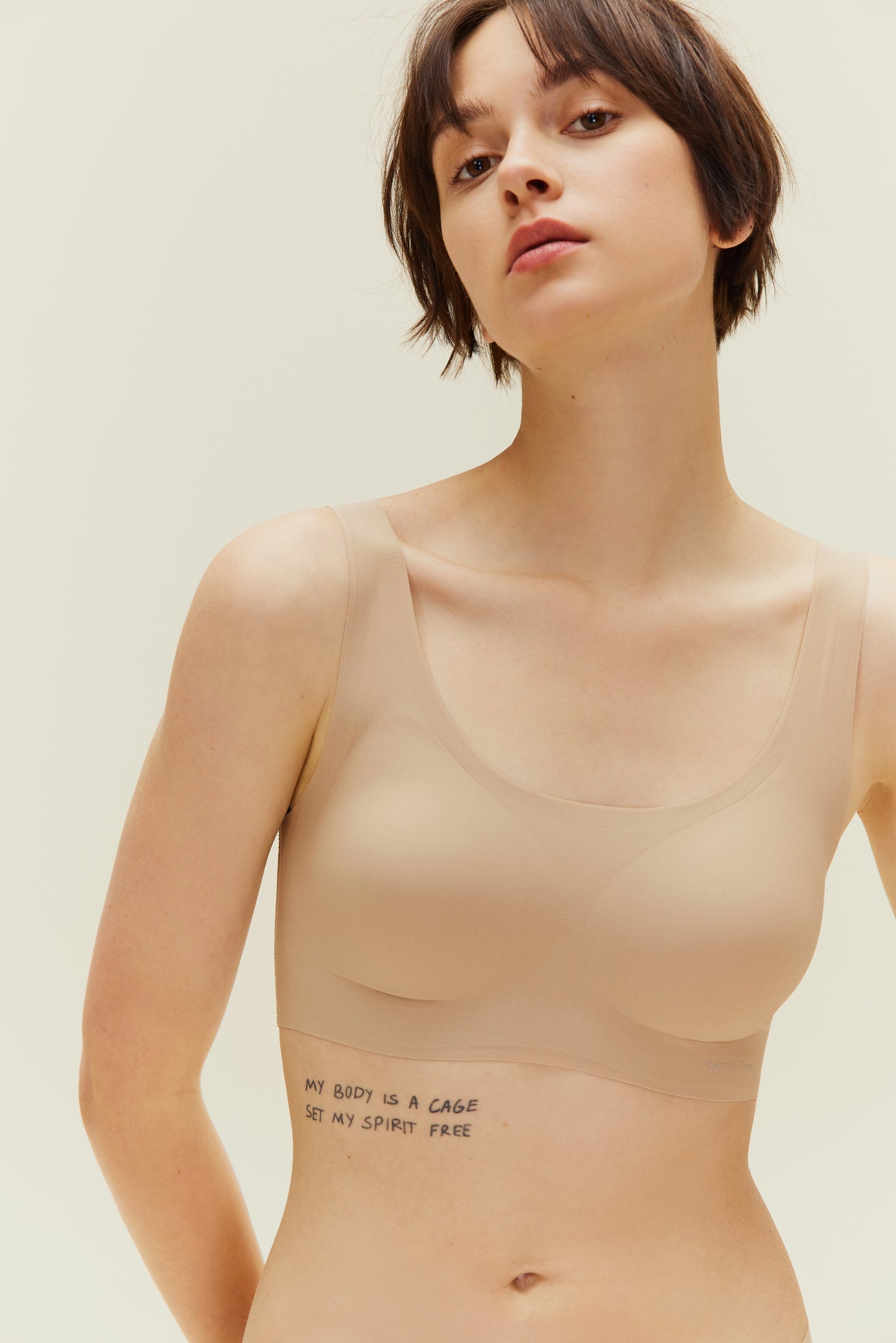 A young woman wearing a Barely Zero Classic Bra Trio in beige, embossed with the phrase "my body is a cage set my spirit free," stands against a light beige background, looking directly at