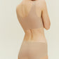 A woman in a beige Barely Zero Classic Bra Bundle bodysuit, viewed from the back, showing the garment's full coverage and a smooth finish across the back and hips. She has short dark hair.