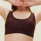 A woman in a dark brown Barely Zero Classic Bra Bundle frames her face with her hands in a diamond shape, focusing on body positivity with a neutral background.