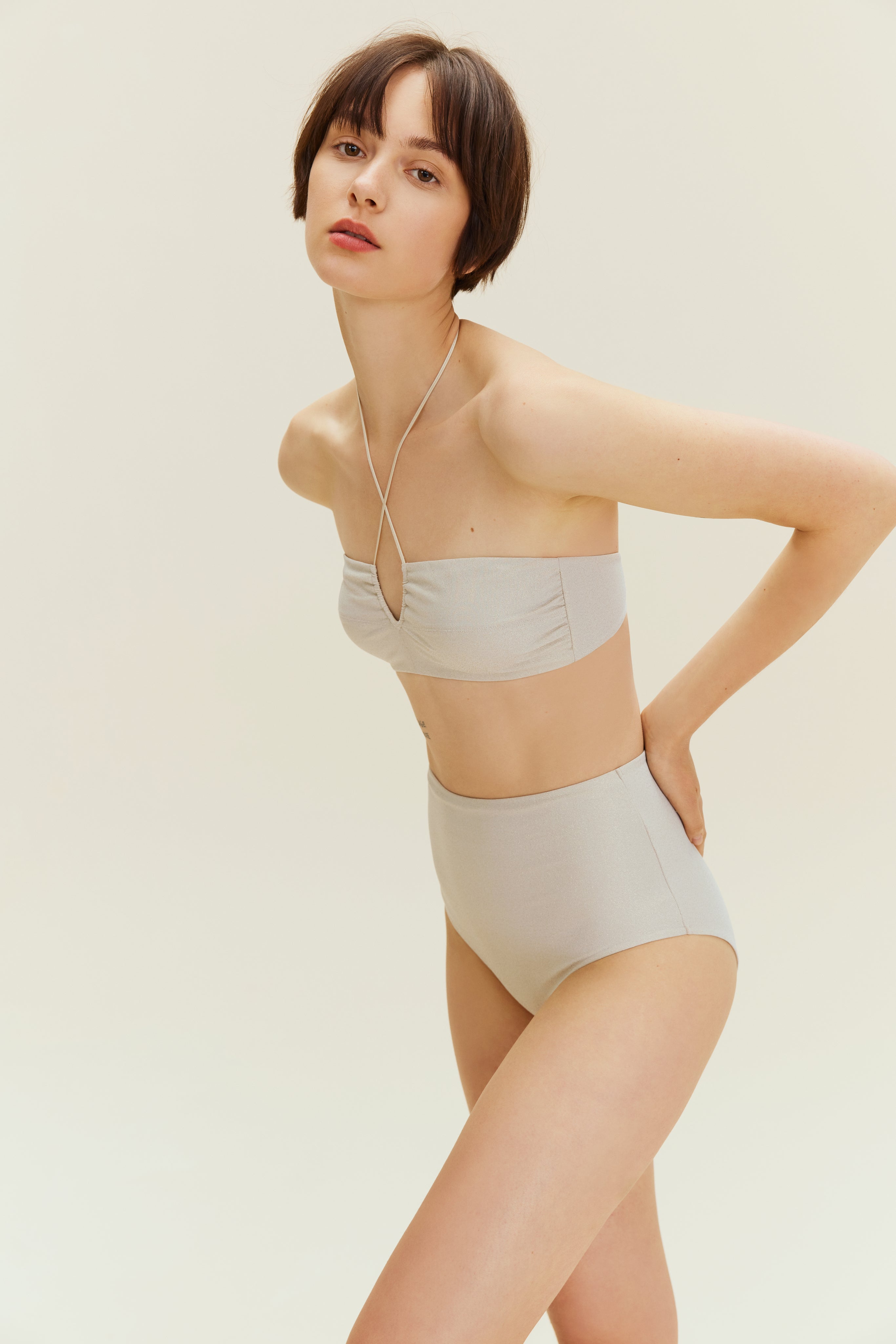 Woman standing leaning forward wearing gold halter neck bikini bop and high waisted bikini bottom