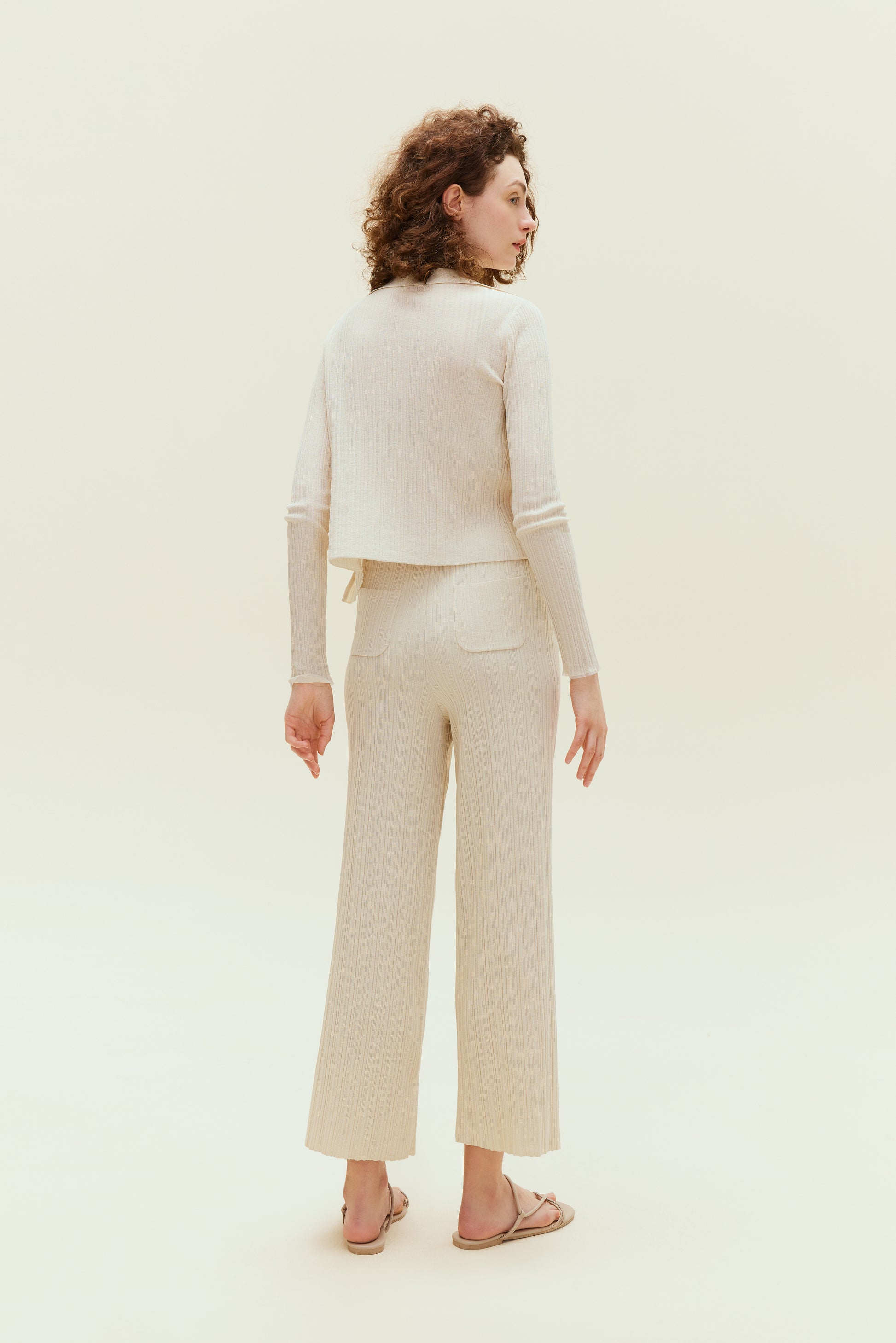 Back view of woman standing wearing off white knit pants with pockets with white knit cardigan