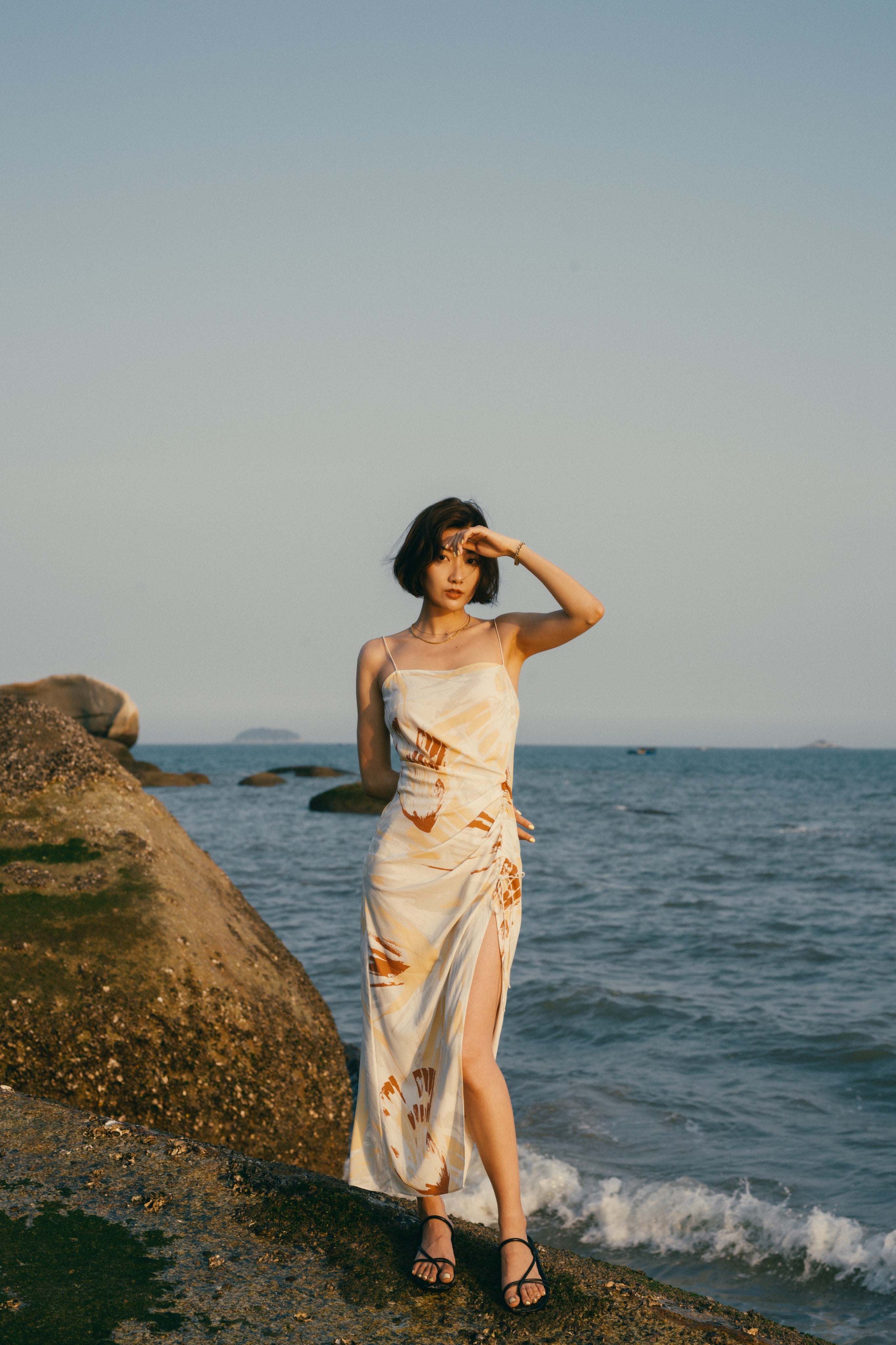 Woman standing on beach wearing spaghetti strap dress with brown, beige, white print with high slit on side