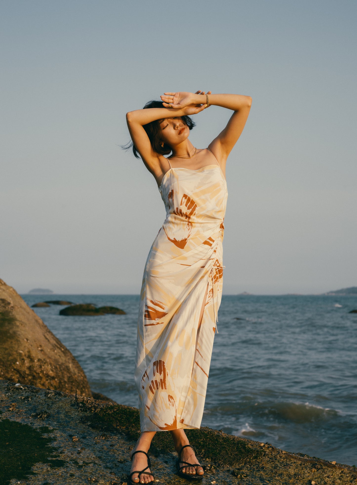 Woman standing on beach wearing spaghetti strap dress with brown, beige, white print with high slit on side