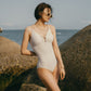 Woman standing by beach wearing gold one piece swimsuit with spaghetti straps and keyhole neckline design