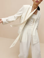 Front view of model wearing ivory blazer with matching color top, ribbon tie, and pants.