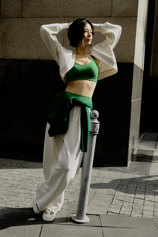 A woman wearing a white sports cardigan, green sports bra, and white sports pants leans on the railing. There is a building in the background.