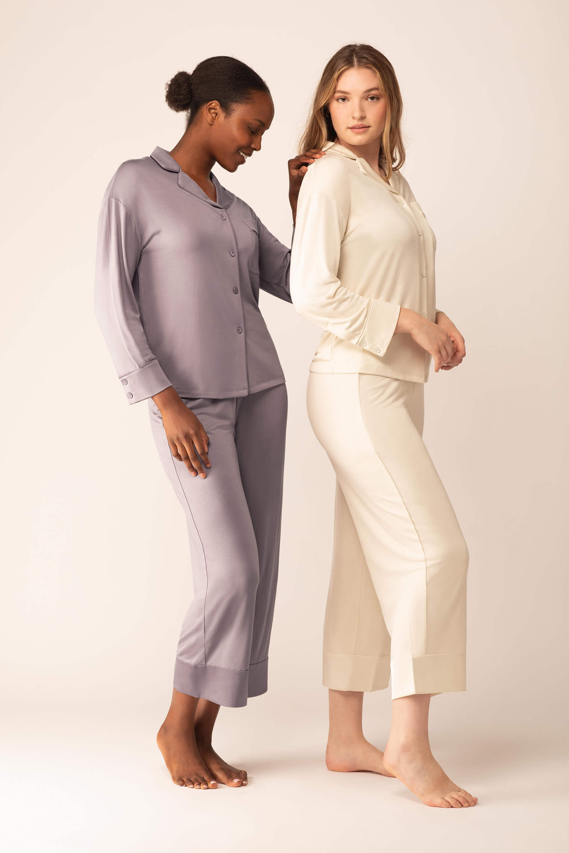 One woman wearing light purple button up pajama shirt with pocket and matching pajama pants next to another woman wearing off-white version of same pajama set