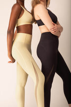 Woman wearing yellow sports bra and leggings with side mesh overlay standing next to woman wearing black sports bra and leggings with side mesh overlay