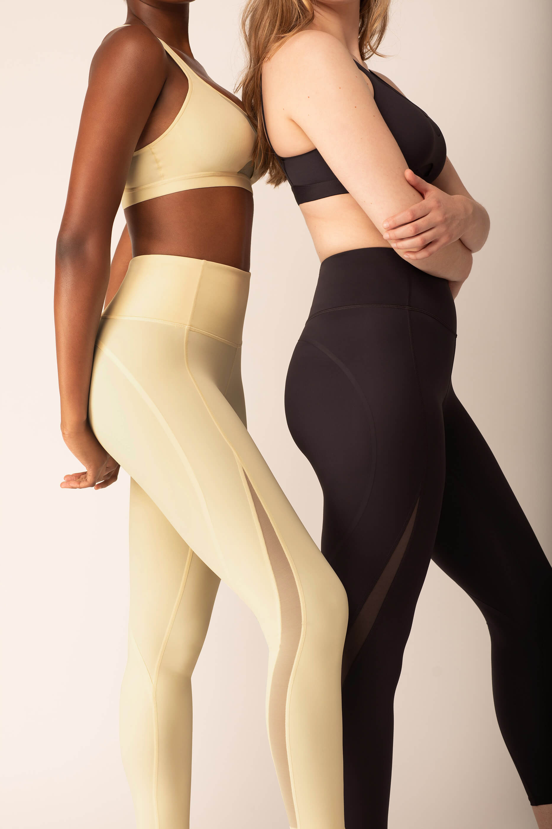 Woman wearing yellow sports bra and leggings with side mesh overlay standing next to woman wearing black sports bra and leggings with side mesh overlay