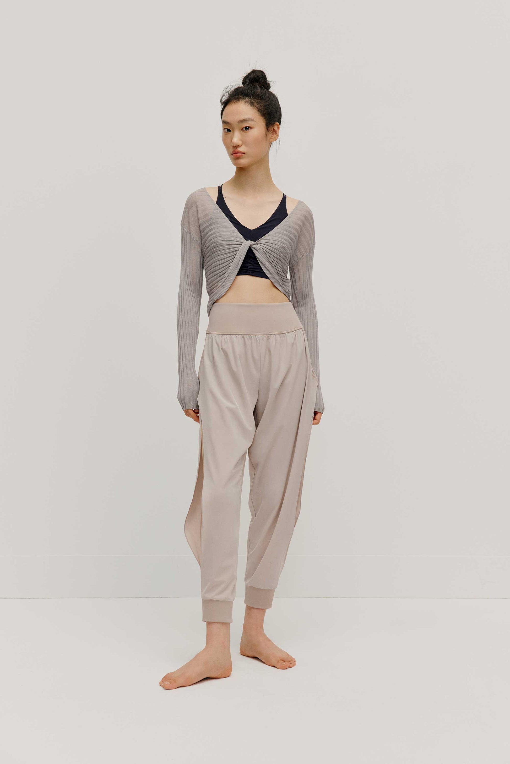 a full body front view of a women wearing a neutral color woven jogger with side slits and a navy bra covered by a mauve color see thru knit top.