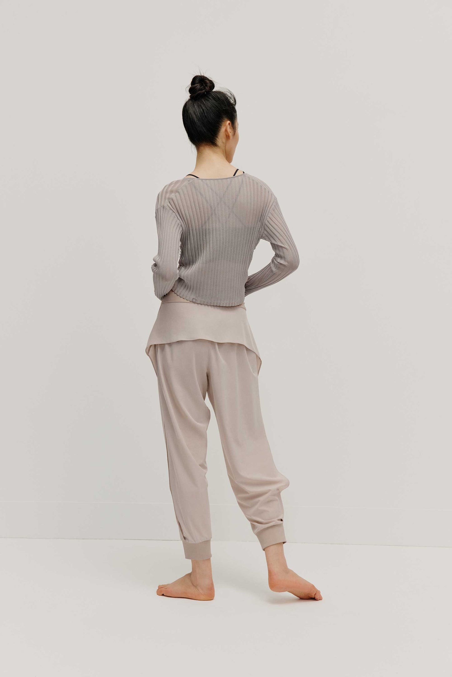 a full body back view of a women wearing a neutral color woven jogger with side slits and a navy bra covered by a mauve color see thru knit top.