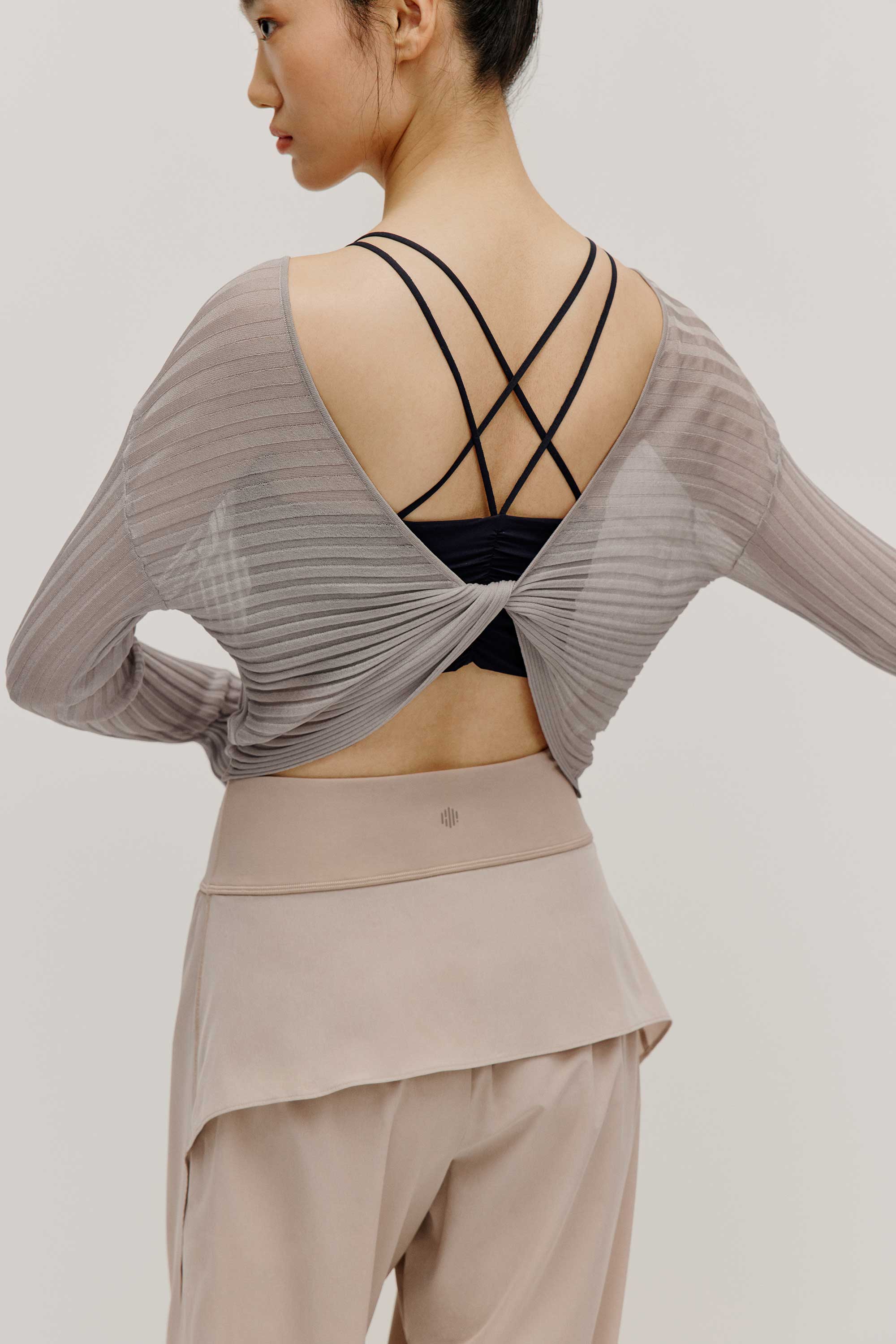 back view of a women wearing a neutral color woven jogger and a navy bra covered by a mauve color see thru knit top with a knot. 