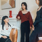 three woman in a living room, two standing and one sitting. Sitting one wearing white satin shirt and black skirt. Middle one standing wearing red cardigan and black pants. Right one standing wearing navy dress