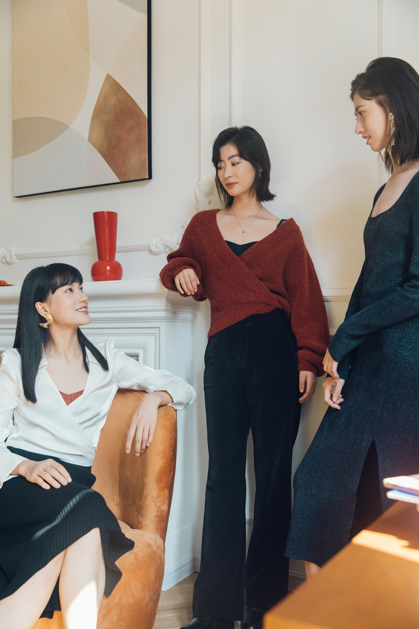 three woman in a living room, two standing and one sitting. Sitting one wearing white satin shirt and black skirt. Middle one standing wearing red cardigan and black pants. Right one standing wearing navy dress