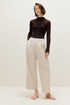 A woman wears a brown silky wool mock neck sweater and white pants.