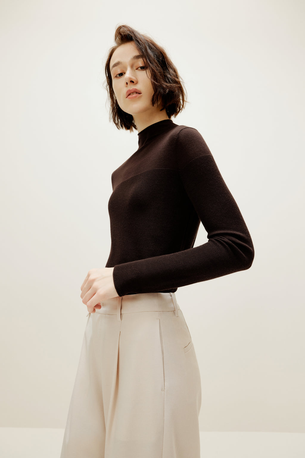 A woman wears a brown silky wool mock neck sweater and white pants.