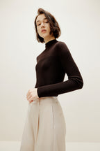 A woman wears a brown silky wool mock neck sweater and white pants.